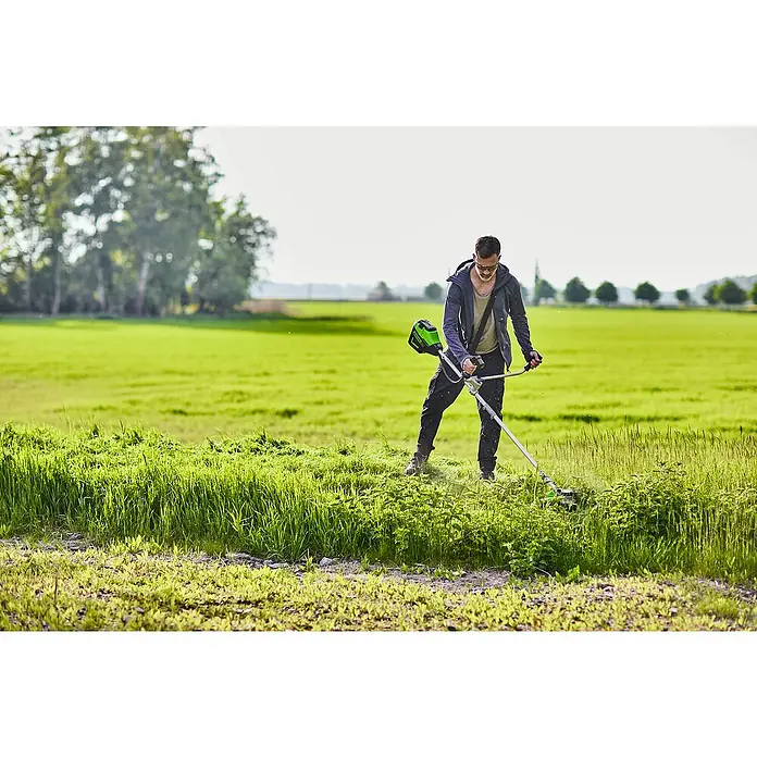 Greenworks 60V Akkusystem Akku-Rasentrimmer GD60BCB - 60 V, Li-Ionen, Ohne Akku, Schnittbreite Messer: 26 cm, Schnittbreite Faden: 40 cm - Ein Mann benutzt einen Rasentrimmer, um das Gras in einem Feld zu schneiden. Der Rasentrimmer ist ein grünes Elektrowerkzeug.