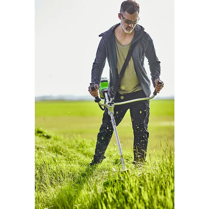 Greenworks 60V Akkusystem Akku-Rasentrimmer GD60BCB - 60 V, Li-Ionen, Ohne Akku, Schnittbreite Messer: 26 cm, Schnittbreite Faden: 40 cm - Mann benutzt einen Rasentrimmer in einem grünen Feld. Der Trimmer ist ein leistungsstarkes Werkzeug für die Gartenarbeit.