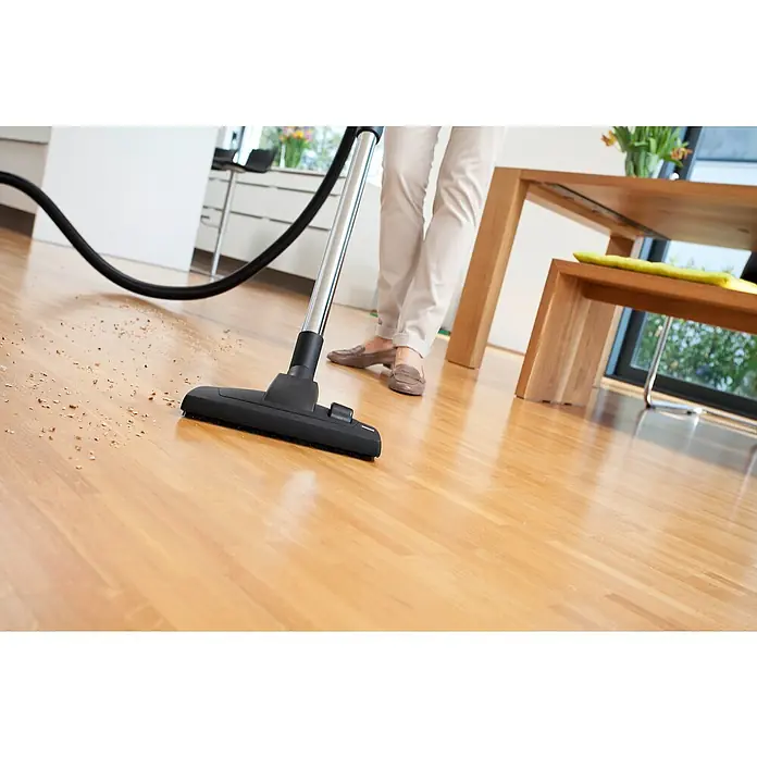 Kärcher Elektro-Staubsauger DS 6 - 650 W - Nahaufnahme eines Staubsaugers, der auf einem Holzboden Krümel aufsaugt, im Hintergrund sind Beine und ein Tisch sichtbar.
