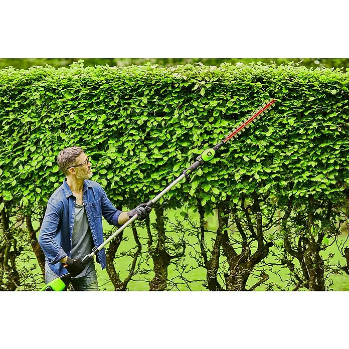 Greenworks 60V Akkusystem Teleskop-Heckenschere GD60PHT51 - 60 V, Li-Ionen, Ohne Akku, Schnittlänge: 51 cm - Ein Mann benutzt eine Heckenschere, um eine hohe grüne Hecke zu schneiden. Die Heckenschere hat einen Teleskopstiel und ist akkubetrieben.
