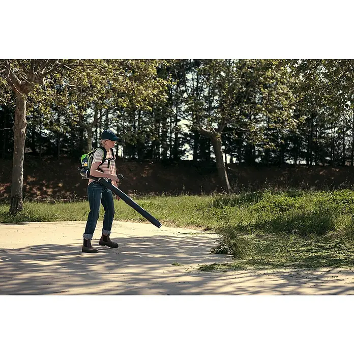 Greenworks 60V Akkusystem Akku-Laubbläser Rucksackbläser GD60BPB - Ohne Akku, Blasgeschwindigkeit: 257 km/h, Blasleistung: 540 m³/min - Eine Person benutzt einen Laubbläser mit Rucksack, um Gras von einem Bürgersteig zu entfernen, mit Bäumen im Hintergrund.