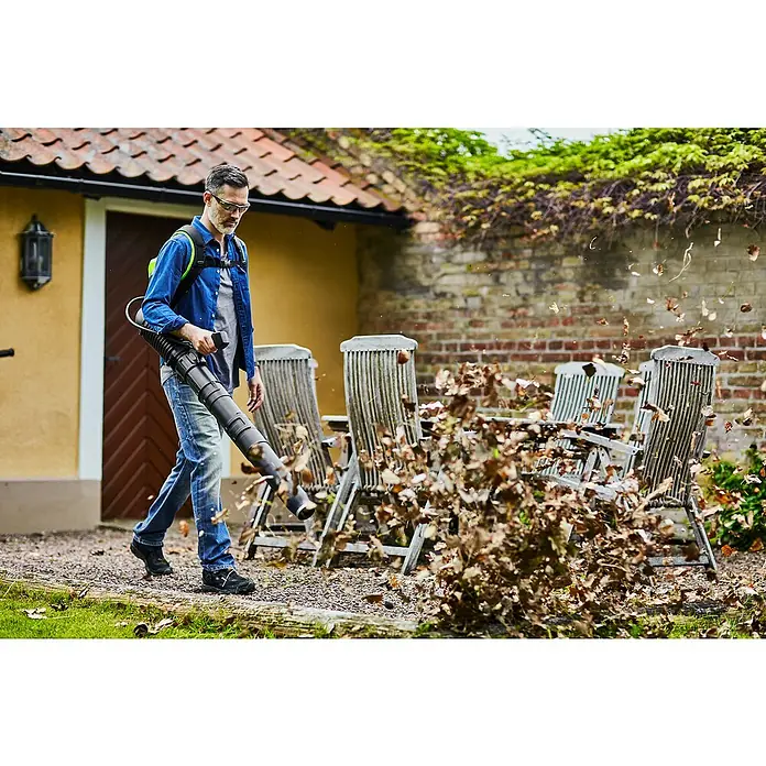 Greenworks 60V Akkusystem Akku-Laubbläser Rucksackbläser GD60BPB - Ohne Akku, Blasgeschwindigkeit: 257 km/h, Blasleistung: 540 m³/min - Ein Mann benutzt einen Laubbläser, um Blätter auf einem Kiesweg vor einem Haus zu entfernen. Der Laubbläser ist der Fokus des Bildes.