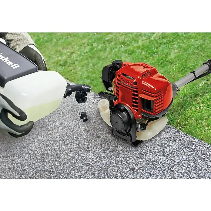 Einhell Desbrozadora de gasolina GC-BC 36-4S - 1 kW, 37,7 cm³, Ancho de corte hilo: 42 cm, Ancho de corte cuchilla: 25 cm - Un trabajador llena de combustible un cortacésped Einhell rojo. El dispositivo está sobre una superficie de hormigón gris con césped al fondo.