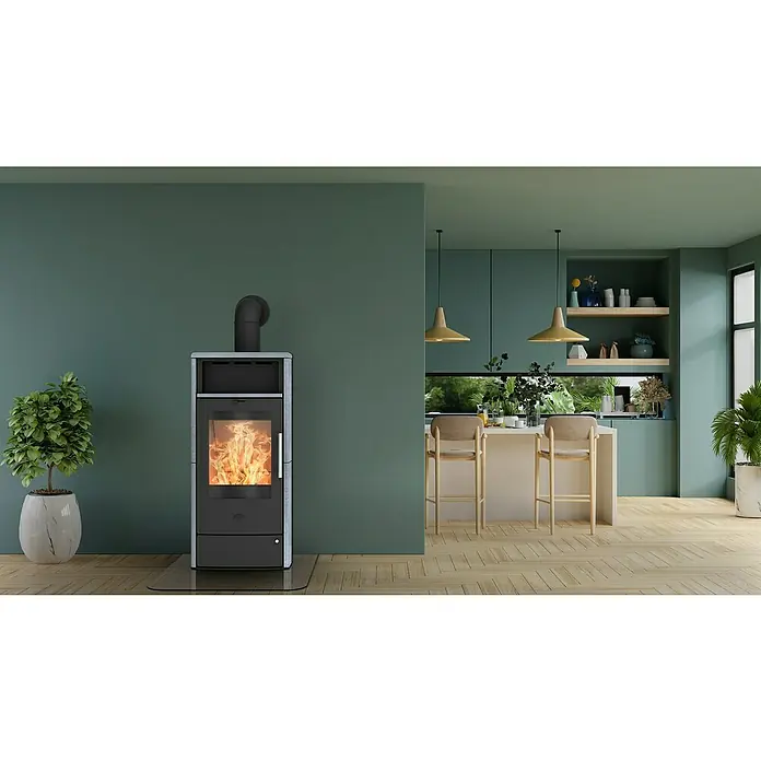 Fireplace Kaminofen Teraga - 6 kW, Raumheizvermögen: 108 m³, Material Verkleidung: Speckstein, Schwarz - Ein Kaminofen mit Glasbodenplatte steht vor einer grünen Wand in einem modernen Raum mit Holzboden. Die Flammen im Ofeninneren leuchten hell.