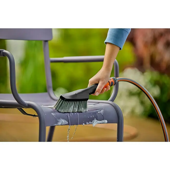 Gardena Komfort-Handbürste Cleansystem S soft - Wasserführend - Eine Person reinigt einen violetten Stuhl im Freien mit einer GARDENA Waschbürste. Die Bürste ist an einen Gartenschlauch angeschlossen.