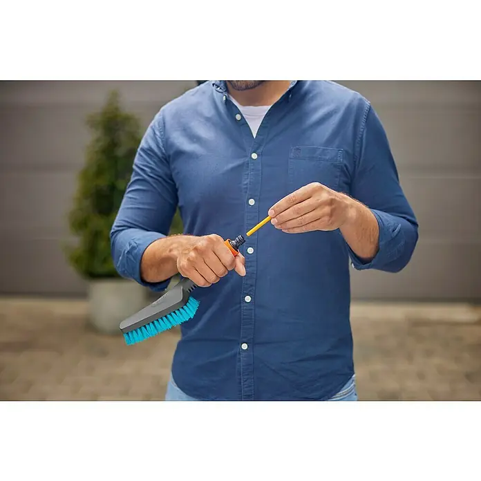 Gardena Handbürste Cleansystem - Regulierbarer Wasserdurchfluss - Ein Mann montiert eine GARDENA Waschbürste an einen Gartenschlauch. Das blaue Bürstenzubehör ist bereit für Reinigungsarbeiten im Freien.