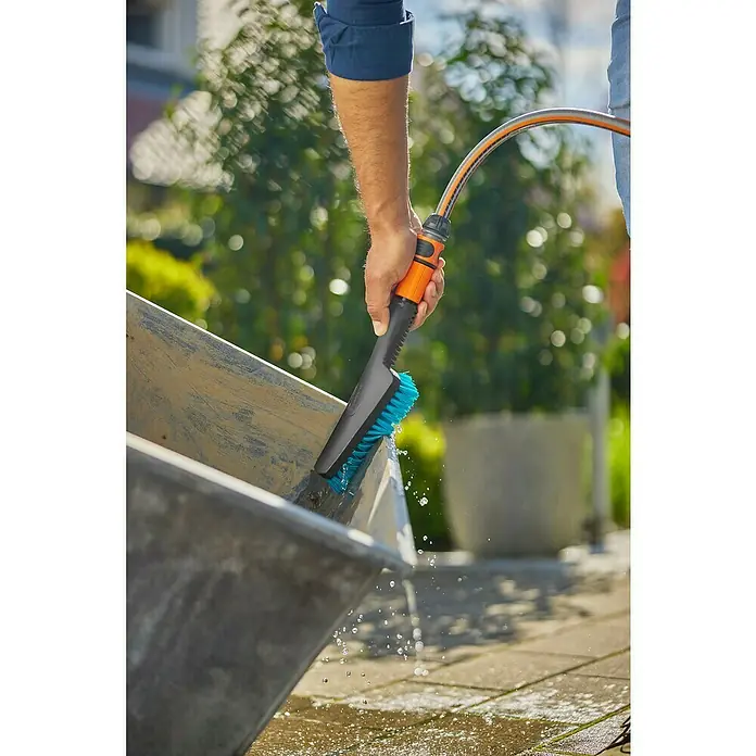 Gardena Handbürste Cleansystem - Regulierbarer Wasserdurchfluss - Der GARDENA Waschbürste mit Wasseranschluss im Einsatz, reinigt eine Schubkarre mit blauen Borsten und Wasserdüsen.