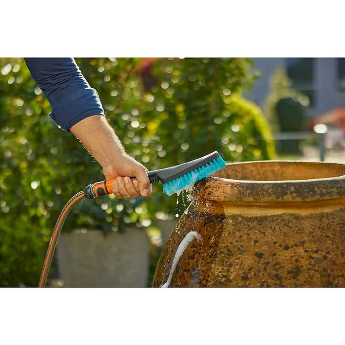 Gardena Handbürste Cleansystem - Regulierbarer Wasserdurchfluss - Eine Person, die einen GARDENA-Scheuerbürste mit Wasseranschluss verwendet, um einen großen Topf im Freien zu reinigen, wobei der Fokus auf der Bürste liegt.