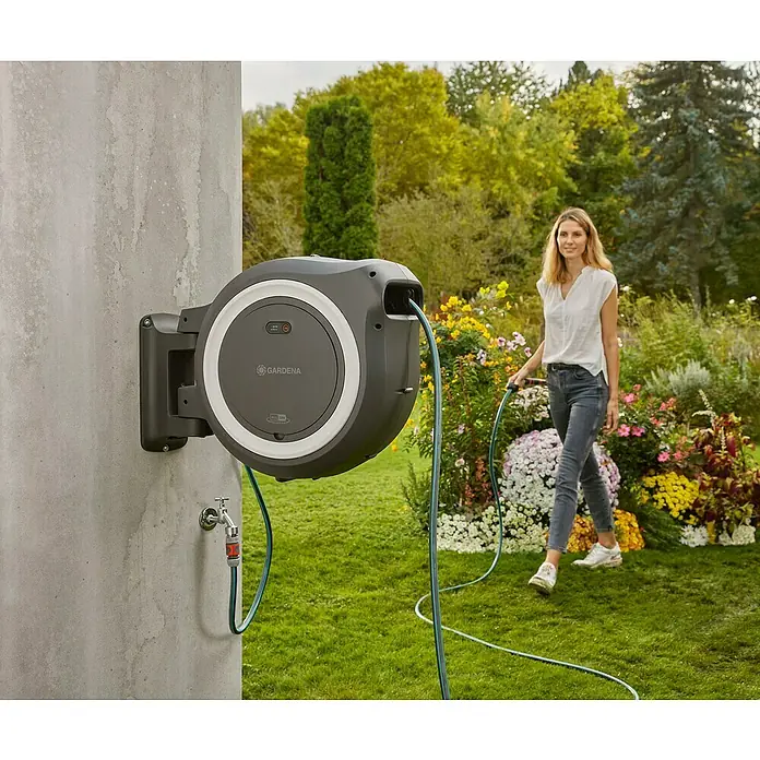 Gardena Power for All 18V Enrollador de manguera de batería Power-Roll XL - 18 V, Sin batería, Longitud del tubo: 35 m - Caja de manguera Gardena gris en una pared con una mujer que sostiene una manguera de jardín en un jardín.