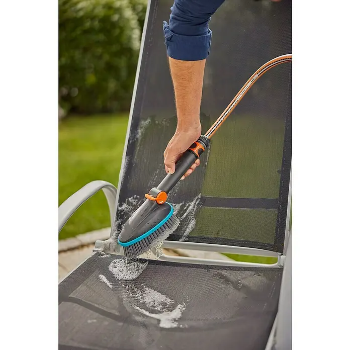 Gardena Cleansystem Komfort-Handbürste Cleansystem M soft - Wasserführend - Eine Person reinigt einen Liegestuhl mit einer GARDENA Reinigungsbürste und einem Wasserschlauch, um Schmutz zu entfernen.