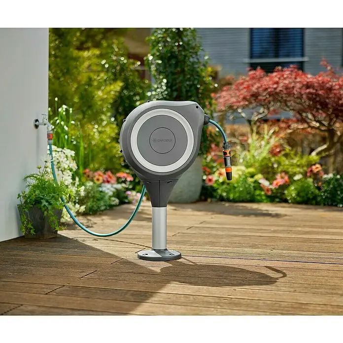 Gardena Schlauchbox Roll-Up M - Schlauchlänge: 20 m, 360 ° drehbar - Das Bild zeigt eine Gardena Schlauchbox an einer Hauswand im Garten. Die Schlauchbox ist grau und hat einen silbernen Standfuß.