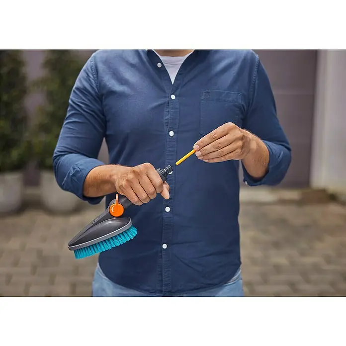 Gardena Cleansystem Handbürste Cleansystem M hard - Wasserführend - Nahaufnahme einer Person, die eine Waschbürste vorbereitet. Die Outdoor-Bürste hat blaue Borsten und einen orangefarbenen Knopf.