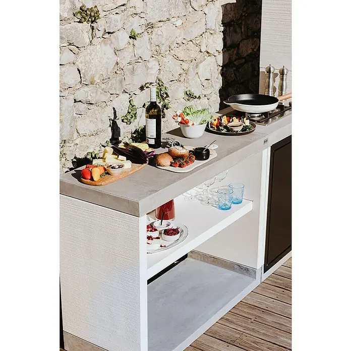 Tuozi Venit Outdoorküchen-Modul Seitenschrank - Weiß-Grau, B x T x H: 110 x 47 x 88 cm - Eine Küchentheke im Freien mit Speisen, Getränken und Kochutensilien auf einer Betonoberfläche, vor einer Steinmauer.