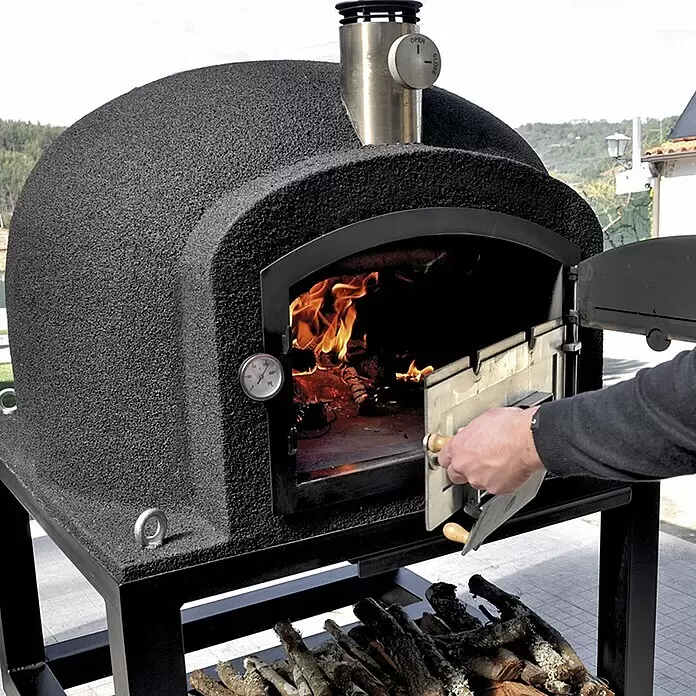 Tuozi Kitaway Outdoorküchen-Modul - Grau/Weiß, B x T x H: 100 x 100 x 65 cm - Schwarzer, texturierter Outdoor-Pizzaofen mit Feuer, auf Ständer mit Holzlager. Eine Person öffnet die Ofentür.