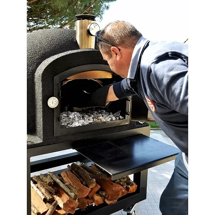 Tuozi Kitaway Outdoorküchen-Modul - Grau/Weiß, B x T x H: 100 x 100 x 65 cm - Mann bedient schwarzen strukturierten Outdoor-Pizzaofen mit Metallschornstein und Thermometer, schürt Kohlen. Brennholz darunter.