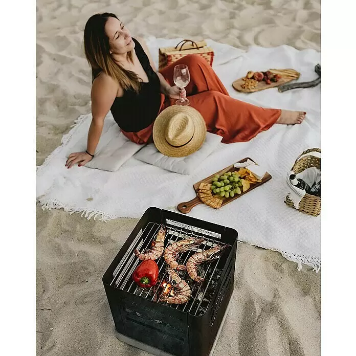 Tuozi Feuerschale Tharsis - L x B x H: 37,5 x 37,5 x 40 cm, Cortenstahl - Schwarzer tragbarer Metallgrill brät Garnelen und Paprika am Sandstrand, mit einer picknickenden Person in der Nähe.