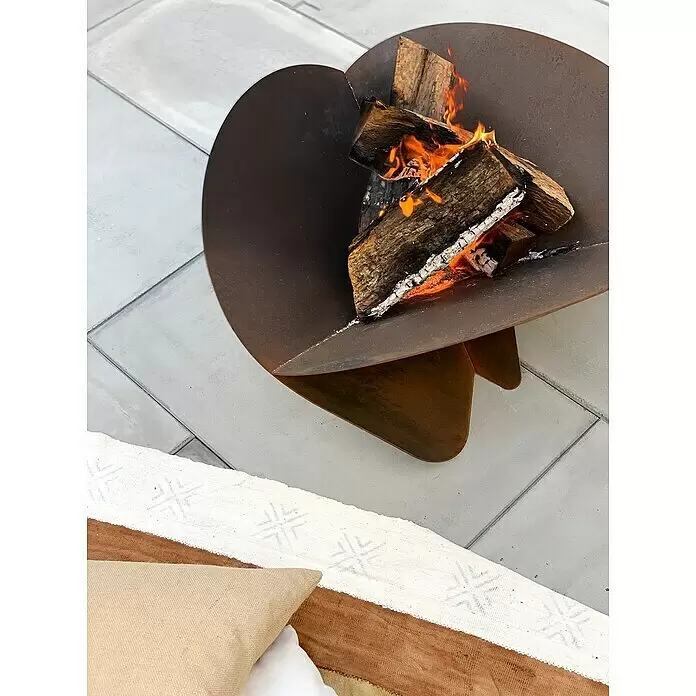 Tuozi Feuerschale Medusae - L x B x H: 75 x 75 x 35 cm, Cortenstahl - Geschwungene Cortenstahl-Feuerstelle mit brennenden Holzscheiten auf einer Terrasse mit Polstern.
