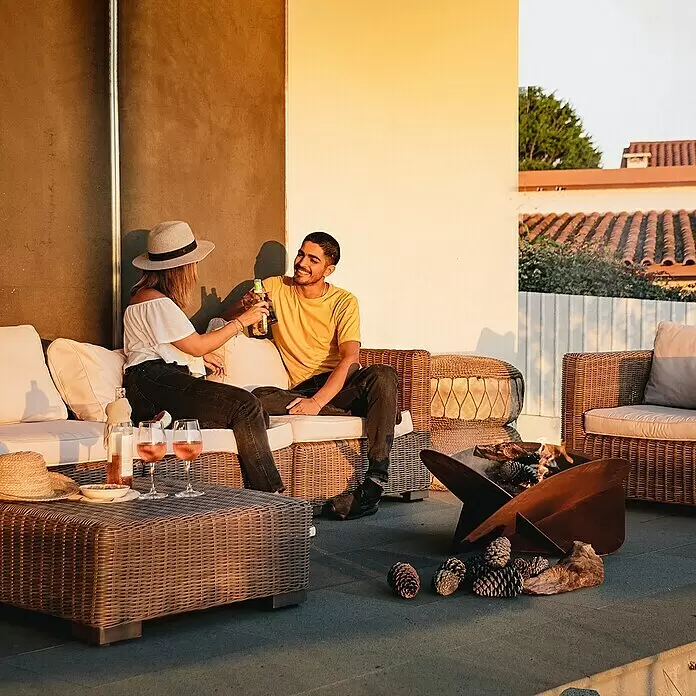 Tuozi Feuerschale Medusae - L x B x H: 75 x 75 x 35 cm, Cortenstahl - Geometrische Rost-Metall-Feuerschale und braune Rattan-Outdoor-Lounge auf Terrasse mit entspanntem Paar.