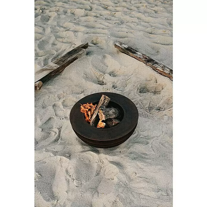 Tuozi Feuerschale Phobos - L x B x H: 60 x 60 x 24 cm, Cortenstahl - Eine dunkle Metall-Feuerschale mit brennenden Holzscheiten steht auf einem Sandstrand, umgeben von Treibholz.