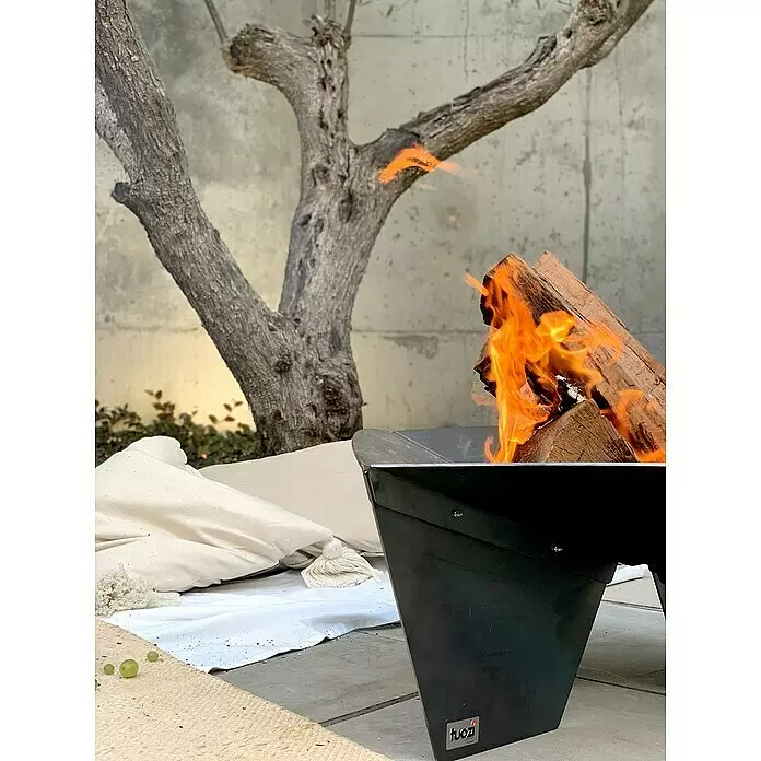 Tuozi Feuerschale Syrtis - L x B x H: 65 x 65 x 31,5 cm, Cortenstahl - Schwarze Metall-Feuerschale von Tuozi mit brennendem Holz, auf einer Terrasse neben Kissen und einem Olivenbaum.