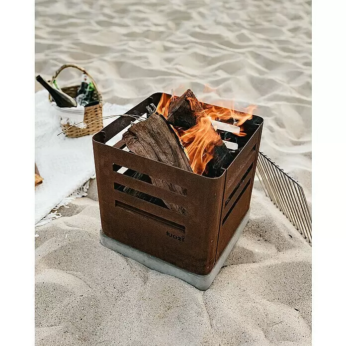 Tuozi Feuerschale Tharsis - L x B x H: 37,5 x 37,5 x 40 cm, Cortenstahl - Eine quadratische, rostbraune Feuerstelle mit Betonsockel, die Holz am Sandstrand verbrennt. Ein Picknickkorb steht daneben.