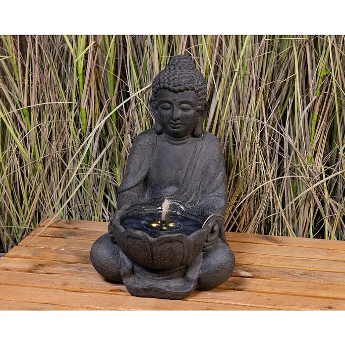 Fuente de jardín LED Mindoro - 28 x 30 x 45 cm - Una estatua de Buda con una fuente de agua en un cuenco de flor de loto, sentada sobre una mesa de madera frente a hierbas.
