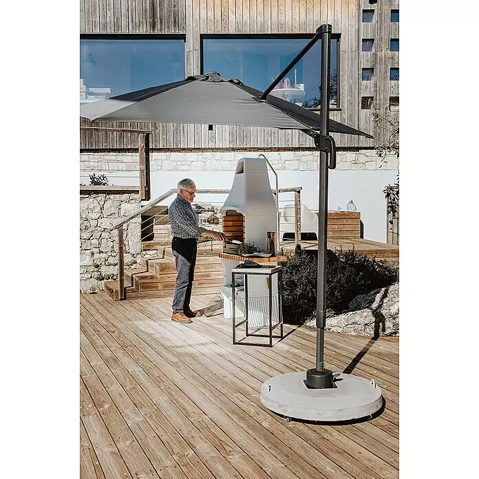 Movelar New Iberia Holzkohlegrill - Grillfläche (B x T): 56 x 35 cm, Grau, B x T x H: 95 x 47 x 190 cm - Großer dunkelgrauer Ampelschirm spendet Schatten auf einer Holzterrasse, wo ein Mann grillt.