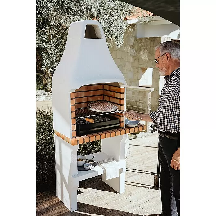 Movelar New Iberia Holzkohlegrill - Grillfläche (B x T): 56 x 35 cm, Grau, B x T x H: 95 x 47 x 190 cm - Mann grillt auf einem weißen Gartengrill mit roten Ziegelakzenten, bereitet Essen über Holzkohle zu.