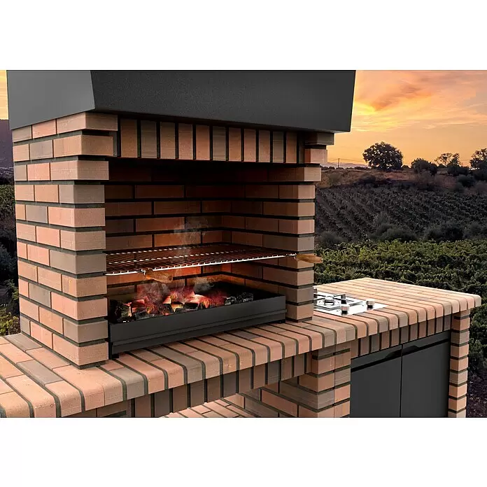 Movelar Volterra Holzkohlegrill - Grillfläche (B x T): 76,5 x 44 cm, Braun - Großer gemauerter Grill mit schwarzer Metallhaube und brennender Kohle, mit einem Gasherd auf der Seitenplatte, vor einer Weinberglandschaft.