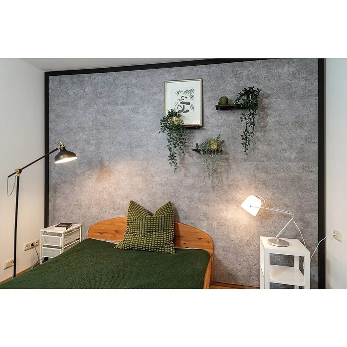 Egger Wandpaneel DecoWall - Rovigo Beton, 125 cm x 66 cm x 12 mm - Ein Schlafzimmer mit einem Bett, einer Stehlampe und einem Nachttischlampe. Das Bett ist mit einer grünen Tagesdecke und einem Kissen ausgestattet.