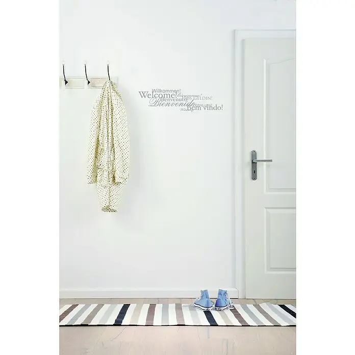 Komar Dekosticker Welcome - 70 x 50 cm, Silber - Helle Eingangshalle mit einer mehrsprachigen Willkommenswandkunst und einem gestreiften Teppich. Die blauen Turnschuhe runden das Bild ab.