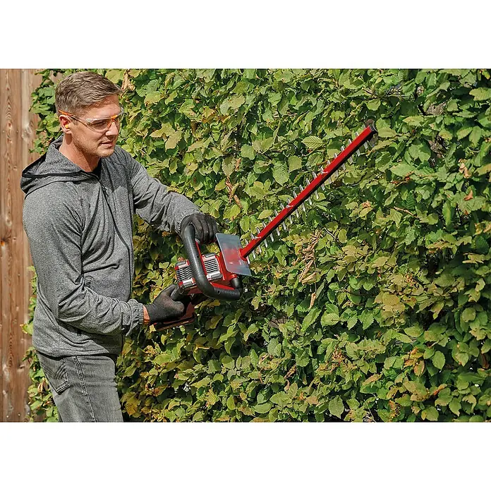 Einhell Power X-Change 18V Akku-Heckenschere GE-CH 36/61 - 18 V, Li-Ionen, Ohne Akku, Schnittlänge: 61 cm - Ein Mann schneidet mit einer roten Heckenschere eine grüne Hecke, Schutzbrille tragend. Die Heckenschere ist das Hauptaugenmerk des Bildes.
