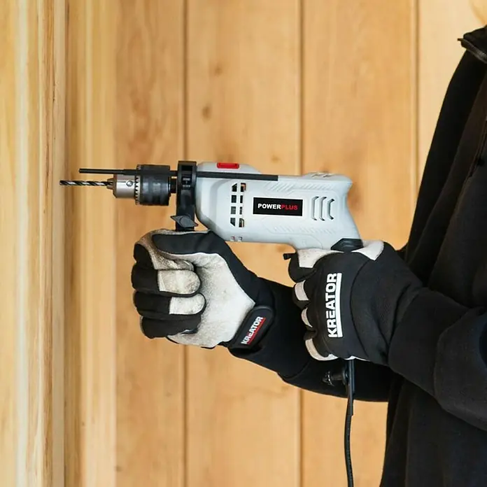 PowerPlus Taladro percutor POWC10110 - 550 W, 3.000 r.p.m., Número máx. de impactos: 4.800 /min - Primer plano de una persona usando un taladro Powerplus para hacer un agujero en una pared de madera, con guantes protectores Kreator.