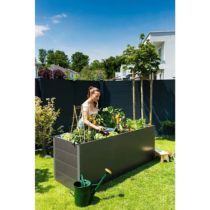 Guardi Hochbeet - 140 x 70 x 76 cm, Metall, Anthrazit - Frau gärtnert in einem grauen Hochbeet mit Pflanzen, Gitter und Gießkanne im Garten unter blauem Himmel.