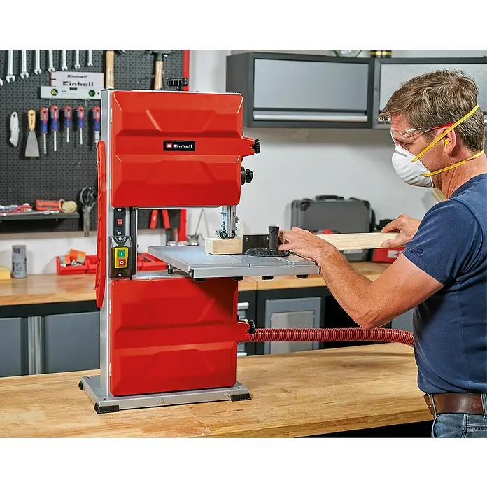 Einhell Sierra de cinta TC-SB 245 L - 400 W, Número de revoluciones de marcha en vacío: 1.450 r.p.m., Longitud de cinta de sierra: 1.712 mm - Un hombre opera una sierra de cinta Einhell roja en un taller, cortando un trozo de madera mientras usa gafas de seguridad y máscara.