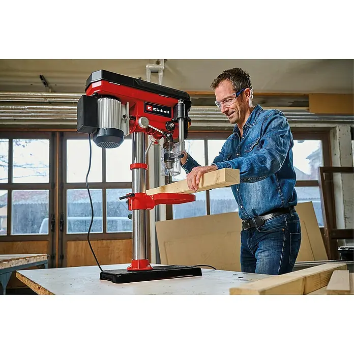 Einhell Taladro de columna TC-BD 630/1 - 630 W, Número de revoluciones de marcha en vacío: 250 r.p.m. - 2.400 r.p.m., Anchura de sujeción mandril portabrocas: 1,5 mm - 16 mm - Un hombre en un taller utiliza un taladro de banco Einhell para trabajar una pieza de madera. Lleva gafas de seguridad.