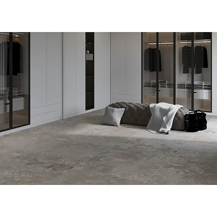 Feinsteinzeugfliese Camarque - 100 x 100 cm, Grau, Matt - Ein begehbarer Kleiderschrank mit grauen Bodenfliesen, einem grauen Tufted Ottoman, einem Kissen, einer Decke und einer schwarzen Handtasche für eine elegante Ästhetik.