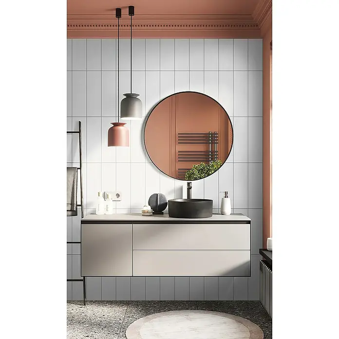 Wandfliese Kaltern - 45 x 15 cm, Weiß, Matt - Badezimmer-Interieur mit einem Unterschrank, einem runden Spiegel und zwei hängenden Pendelleuchten in Rosa und Grau.
