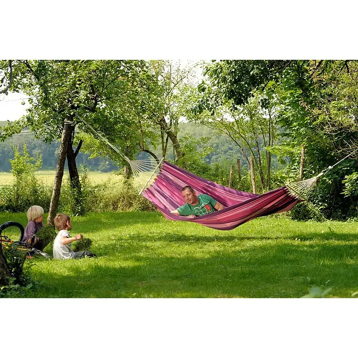 Amazonas Stabhängematte Tonga Candy - L x B: 310 x 100 cm, Rosa/Violett - Mann entspannt in einer gestreiften Hängematte im Garten, während Kinder im Gras spielen. Eine idyllische Szene.