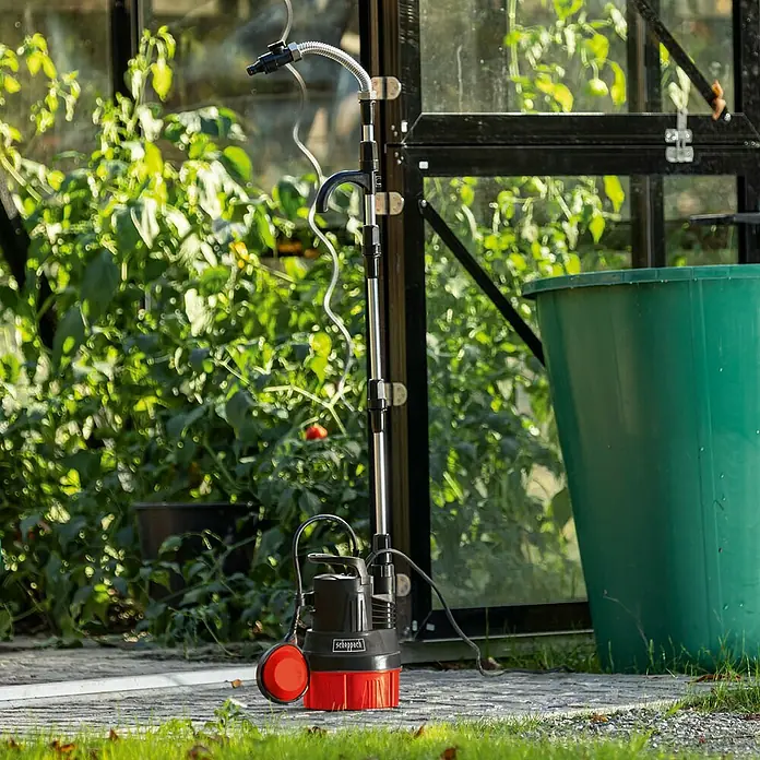 Scheppach Regenfasspumpe RBP350 - 350 W, Max. Fördermenge: 5.500 l/h - Eine Scheppach Tauchpumpe steht vor einem Gewächshaus. Die Pumpe ist schwarz und rot und hat einen silbernen Auslauf.