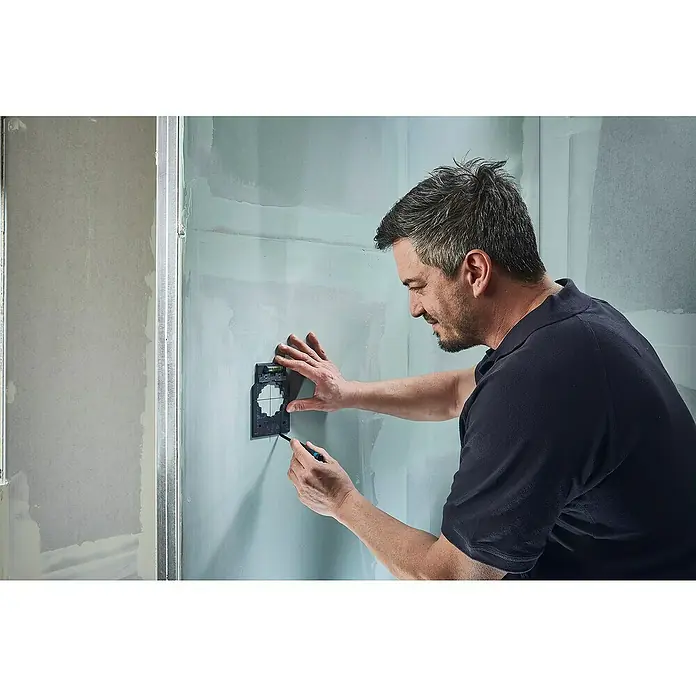 Wolfcraft Kit de accesorios - 3 pzs., Específico para: Cajas de enchufes en pared hueca - Un artesano monta una caja de conexiones negra con nivel de burbuja en una pared para marcar con precisión la instalación eléctrica.