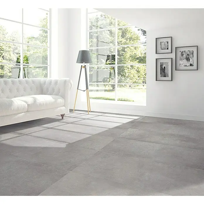 Feinsteinzeugfliese Rimini - 60 x 60 cm, Grigio, Matt - Eine moderne Wohnraumansicht mit grauem Fliesenboden, weißem Sofa und Stehlampe neben einem großen Fenster.