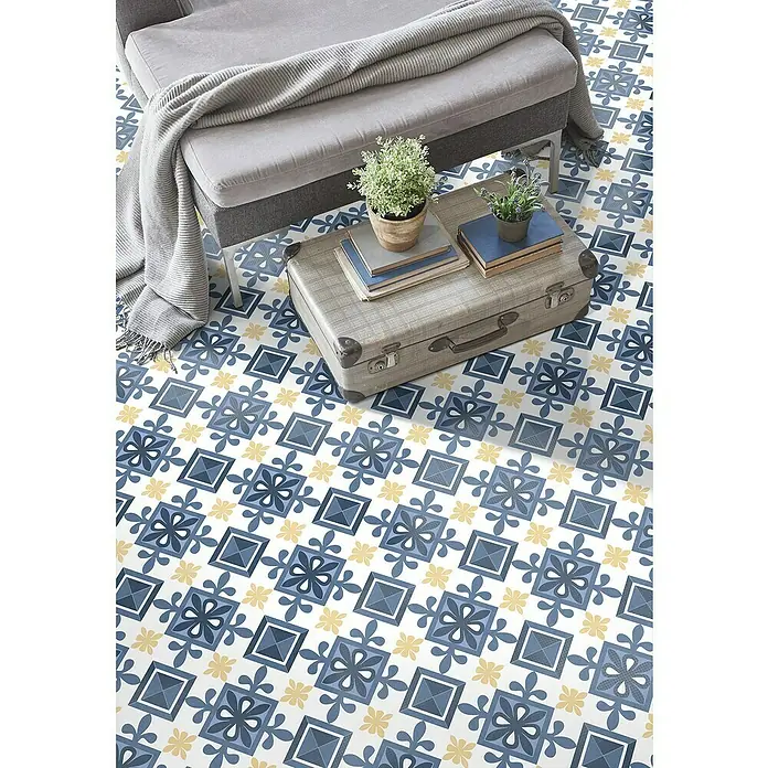 Feinsteinzeugfliese Cordoba Alcazar - 24,8 x 24,8 cm, Bunt, Matt - Elegante Bodengestaltung: Ein Vintage-Koffer dient als Beistelltisch mit Pflanzen und Büchern, stilvoll ergänzt durch ein blau-gelbes Fliesenmuster.