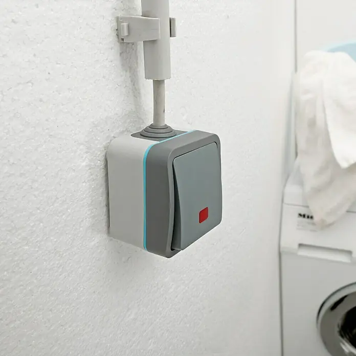 Voltomat Blueline Feuchtraum-Kontrollschalter - Grau, Aufputzmontage, IP54 - Nahaufnahme eines grauen Lichtschalters mit blauen Akzenten an einer weißen Wand, der eine moderne und funktionale Ästhetik bietet.