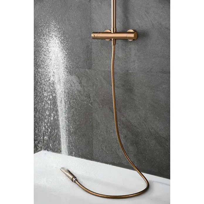 Imex Nápoles Sistema de ducha - Grifo termostático, Oro rosa - Moderno juego de ducha en tonos dorados con mezclador termostático y teleducha bajo el agua corriente contra una pared de piedra gris.