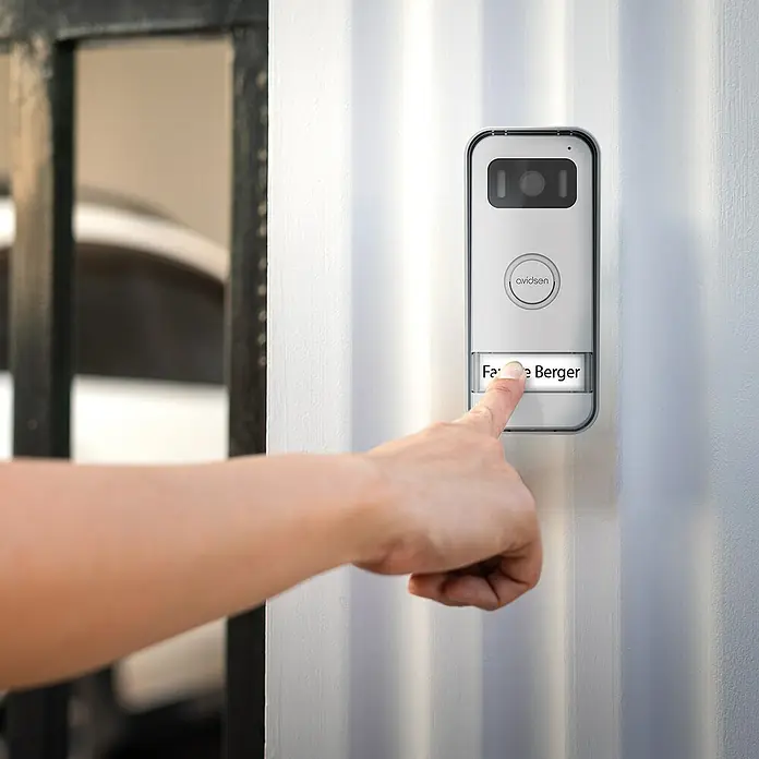 Avidsen Türklingel mit Kamera Smarte Video Türsprechanlage Elia - Weiß, Silber, 800 x 480 Pixel - Eine Hand drückt auf die Klingel, eine moderne Avidsen Türklingel mit Kamera und Namensschild. Sie ist an einer weißen Wand montiert.