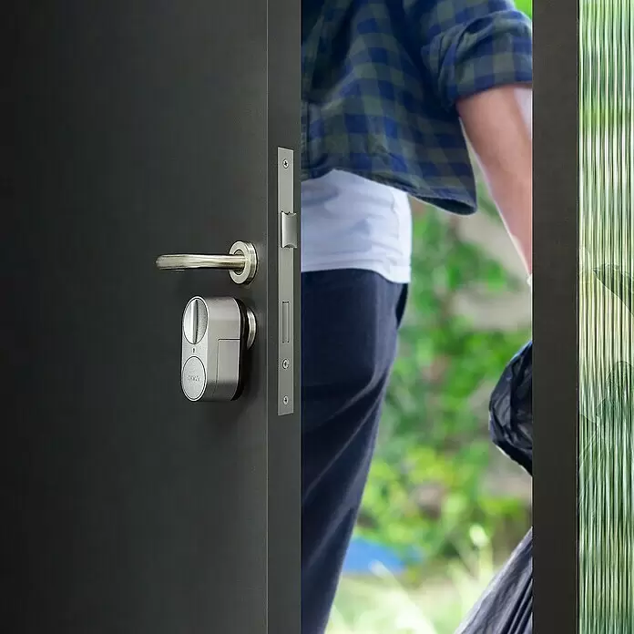 Set de cerradura electrónica Retrofit DiY - Gris, Número de componentes: 3 ud. - Cerradura inteligente gris plateada en una puerta oscura, vista mientras una persona sale por la entrada.