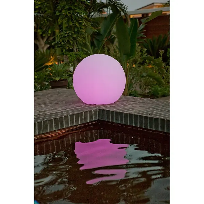 Starlux Bola solar Venus 30 - 0,5 W, 30 cm, RGB, 50 lm - Orbe iluminado: Un orbe rosa iluminado se sienta en una pared de ladrillo junto a un estanque, proyectando una luz colorida.