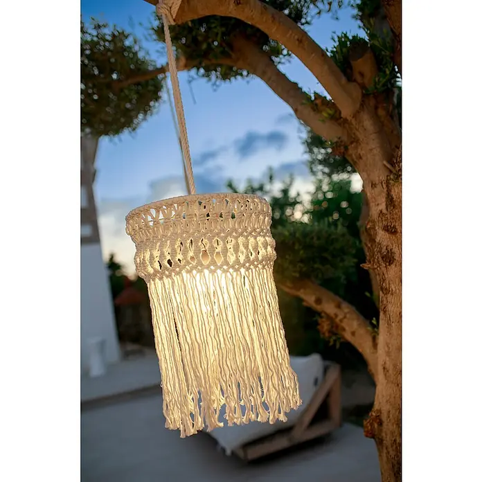 New Garden Lámpara colgante Mykonos - 9 W, Ø x Al: 30 x 40 cm, Blanco - Primer plano de una linterna de macramé para exteriores, suspendida, que irradia un brillo cálido, con la rama y el entorno borrosos al fondo.