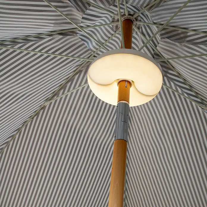 New Garden Plafón LED para parasol Lima - Ø x Al: 17,5 x 6,75 cm - Una vista hacia arriba captura una sombrilla elegante con iluminación integrada a lo largo del poste de madera. El patrón de rayas añade un toque elegante.